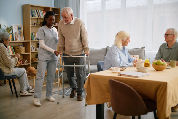 Elderly man using walker assisted by caregiver in care facility with other seniors in background.