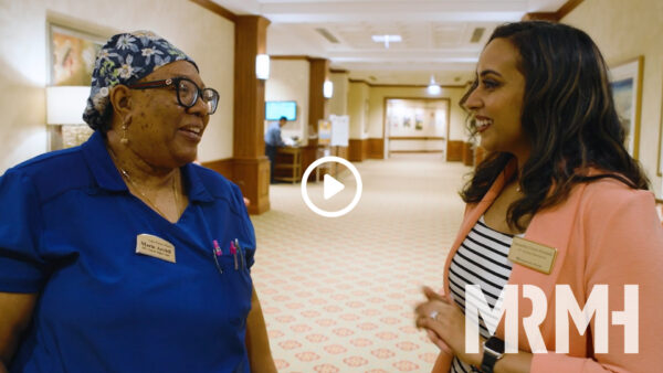 Smiling registered nurse wearing glasses and a blue shirt speaking with a smiling woman in common area of healthcare facility.