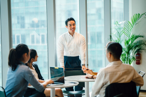 Man standing with large office windows behind him participating in a discussion with four colleagues seated in front of him.