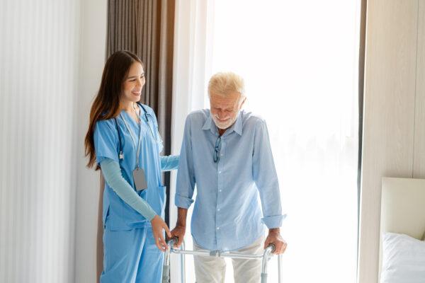 Female care worker in blue scrubs helping an elderly man walk with a walker.